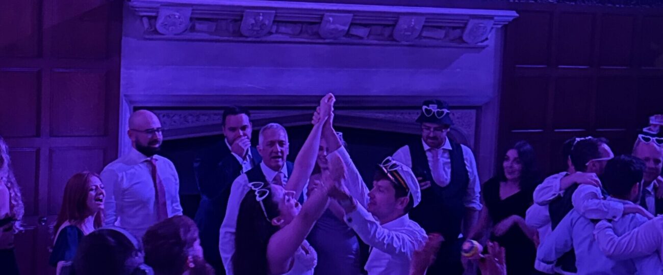 Wedding Reception Guests Celebrating | Mr Beats Wedding guests celebrating with raised hands on the dancefloor under purple DJ lighting
